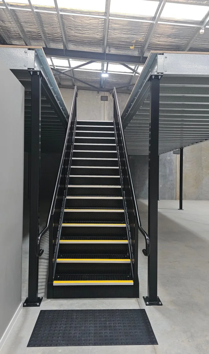 Steel access stair with yellow tread nosings rising to a mezzanine deck inside a warehouse